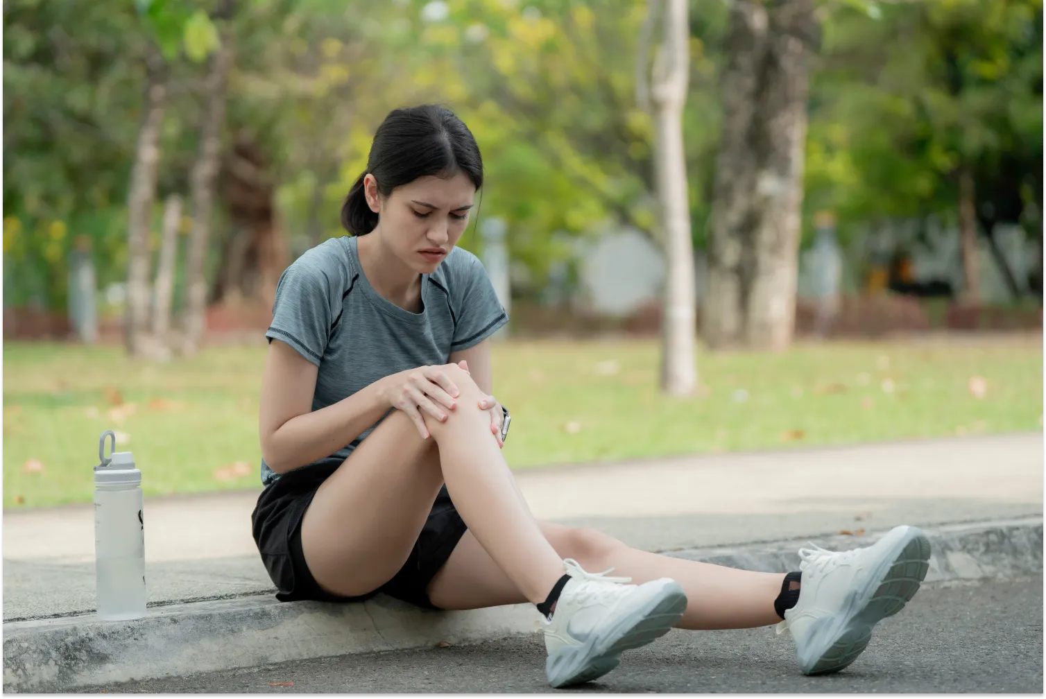 Woman sitting on a curb outdoors holding her knee in pain after exercise — common reasons why your knee might be hurting during or after physical activity product